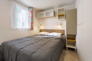Un dormitorio con cama, ventana con cortinas translúcidas, estanterías empotradas e iluminación en la pared.