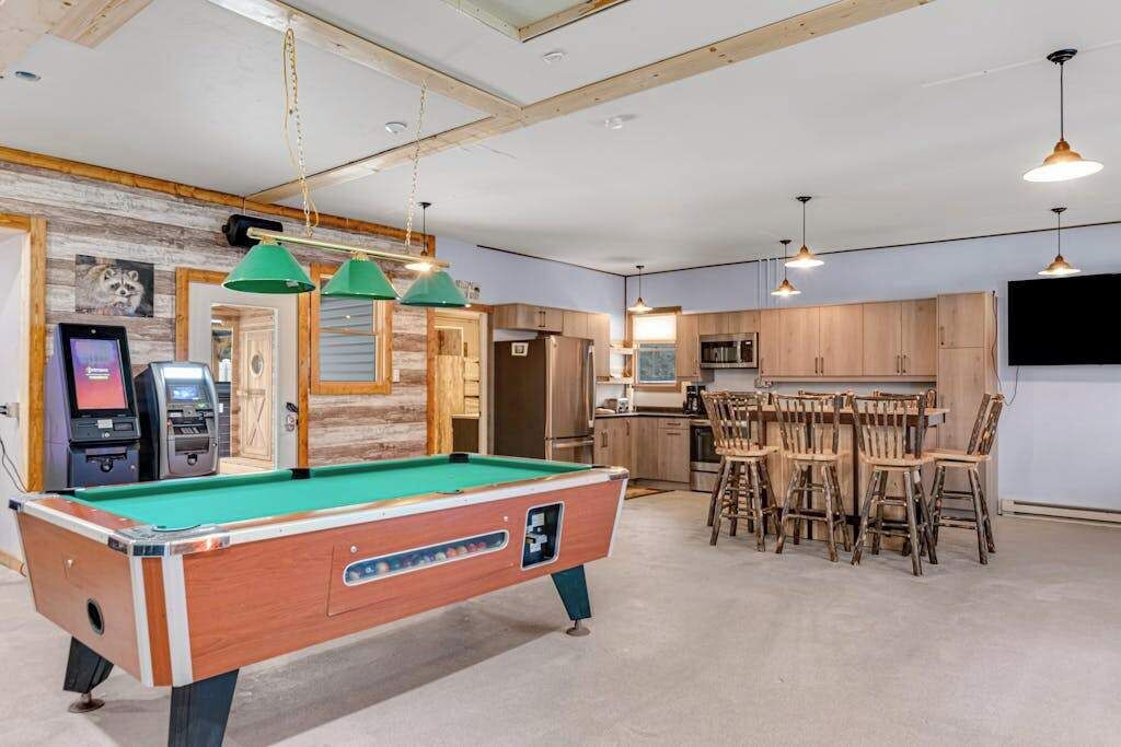 A pool table in a room with wooden cabinets, a bar area with stools, and a television mounted on the wall.