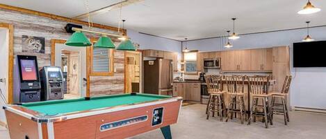 A pool table in a room with wooden cabinets, a bar area with stools, and a television mounted on the wall.