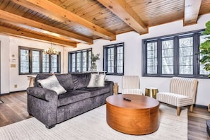 A spacious living room with a dark grey sofa, a round wooden coffee table, and a striped armchair. Large windows provide natural light and a view of trees outside.