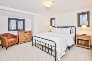 Un dormitorio con cama, una silla, tocador, dos mesitas de noche y dos ventanas.