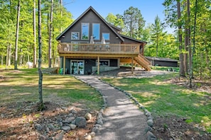 A two-story cabin with a deck, surrounded by trees. - Gorgeous lake home with private pier, beach, firepit, deck, & grill + water toys (Eagle River)
