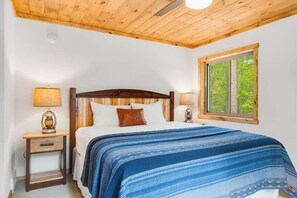 Un dormitorio con techo de madera, una cama con colcha azul estampada, dos lámparas de noche y una ventana con vistas a árboles.