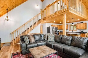 A spacious living area with a large leather sofa, a wooden staircase, and a kitchen in the background. - Gorgeous lake home with private pier, beach, firepit, deck, & grill + water toys (Eagle River)