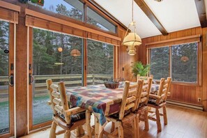 Un comedor de madera con una mesa, sillas, ventanas amplias y una lámpara colgante.