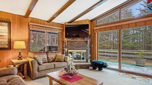 A cozy living room with a fireplace, wooden walls, and large windows. - Lakefront Home with a Private Dock, Deck, Fireplace, Spacious Yard, & Playset (Eagle River)