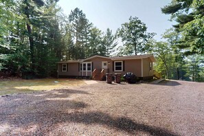 A two-story house with a front porch, a gravel driveway, and a wooded backdrop. - Waterfront Thoro Cottage on Minocqua Chain | Screened Porch, Firepit & Lake Fun! (Minocqua)