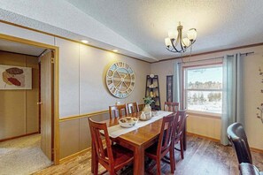 Ein Esszimmer mit Holztisch, Stühlen, einer großen Wanduhr, einem Kronleuchter und einem Fenster mit Vorhängen.