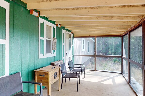 A covered porch with a wooden table and chairs, a small cabinet, and a window with a view of a house and trees.