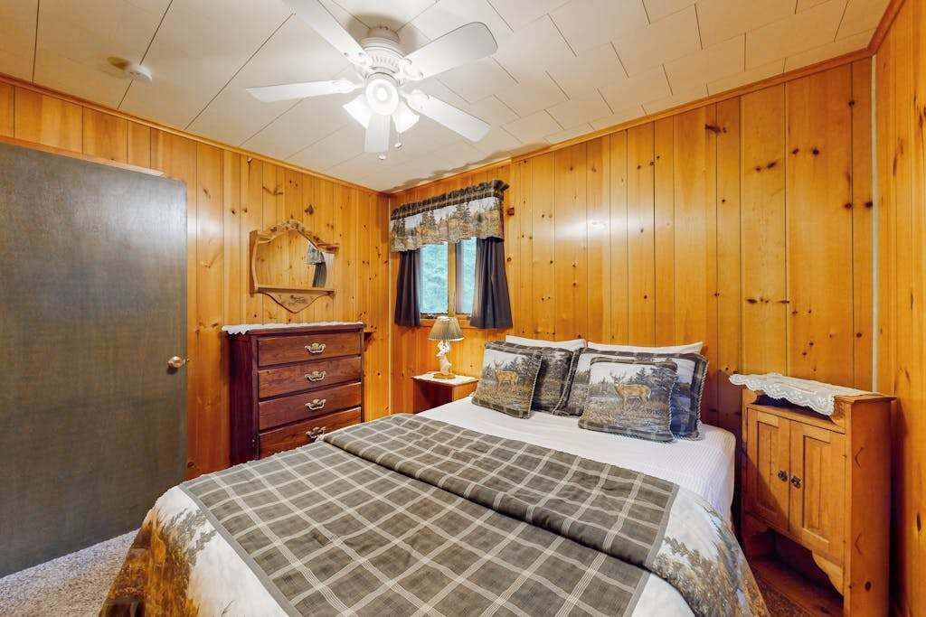 A bedroom with wooden walls, a bed with plaid bedding, a ceiling fan, a dresser, and a window with curtains.