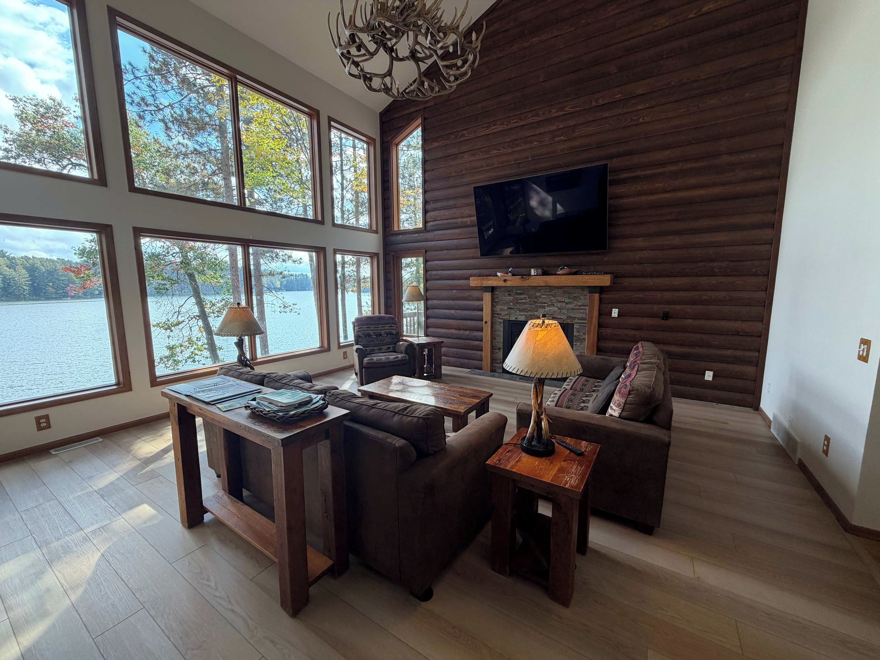 Amplia sala de estar con chimenea, ventanas grandes con vistas a un lago y un televisor montado en la pared.