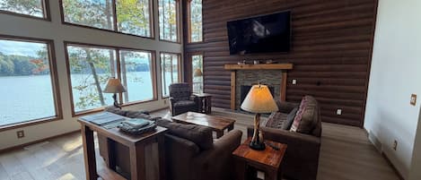 A spacious living room with a fireplace, large windows overlooking a lake, and a television mounted on the wall.