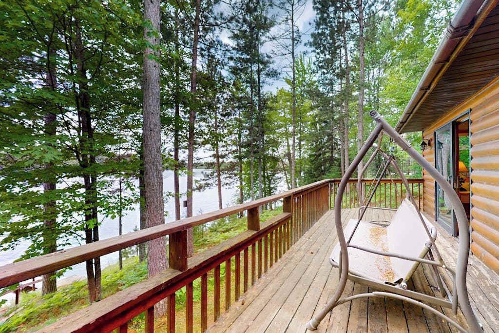 A wooden deck with a metal swing chair overlooking a lake surrounded by trees.