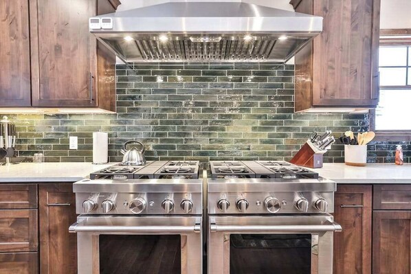 A kitchen with two stainless steel stoves, a large range hood, and a green-tiled backsplash.