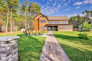 A two-story house with a brick exterior, a stone pathway leading to a patio with outdoor furniture, and a forested backdrop.