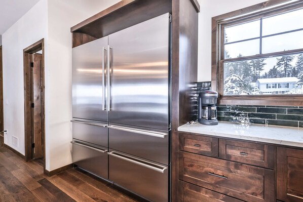 A stainless steel refrigerator, wooden cabinets, and a window with a view of trees and a house. - Dog-friendly lakefront home with firepit, 2 grills, fireplace, dock, & kayaks (St. Germain)
