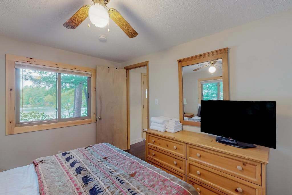 Un dormitorio con cama, cómoda, televisor, ventilador de techo y ventana.