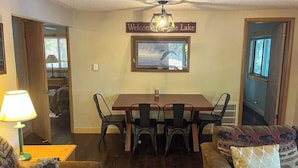 Ein Essbereich mit einem Holztisch, Stühlen, einem Schild mit der Aufschrift 'Welcome to Lake', einem gerahmten Bild von einem Vogel und einer Couch mit gemusterten Kissen.
