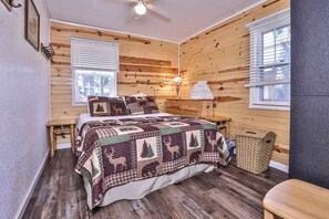 A cozy bedroom with a wooden wall, a bed with a patterned quilt, a bedside lamp, and a window with blinds. - Lake view, dog-friendly cabin with porch & shared dock, swimming area, firepit (St. Germain)