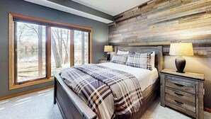 A bedroom with a large window, a bed with plaid bedding, a nightstand with a lamp, and a wooden accent wall. - Dog-friendly lodging w/ scenic deck, pool table, lake dock, & free WiFi (St. Germain)