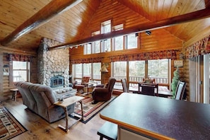 A log cabin interior with a stone fireplace, wooden ceiling beams, and a dining area with a view of the outdoors. - Spacious and beautiful lakefront log home on 7 acres with private dock (Presque Isle)