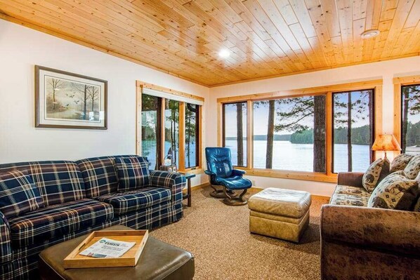 A cabin interior with a plaid sofa, a recliner, a wooden ceiling, and large windows overlooking a lake.