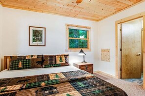 A bedroom with a wooden ceiling, a bed with a quilt, a nightstand, a window, and a door.