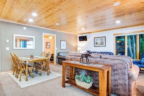 A cozy living room with a wooden ceiling, a dining table with chairs, a sofa, and a television on the wall.