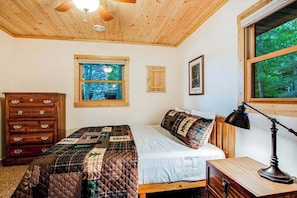 A bedroom with a wooden ceiling, a bed with a quilt, a dresser, a window with a view of trees, and a framed picture on the wall.