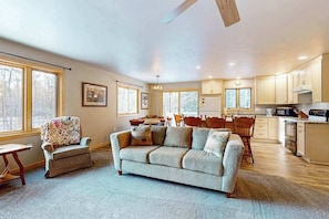 A spacious living area with a sofa, a dining table, and a kitchen in the background. - Cozy tree-lined home located near town, lakes, walking & bike trails, w/ firepit (Manitowish Waters)