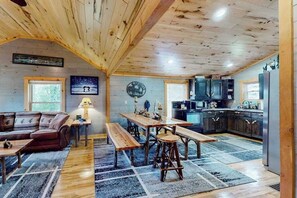 A cabin interior with a wooden ceiling, a dining area with benches, a sofa, a kitchen with a refrigerator, and a window with a view of trees. - Main Level Cozy Home, Conveniently located near downtown by Lakes & Trails. (Mercer)