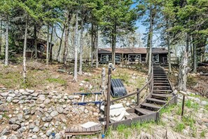 A rustic cabin with a wooden staircase leading to a deck, surrounded by trees and a stone wall.