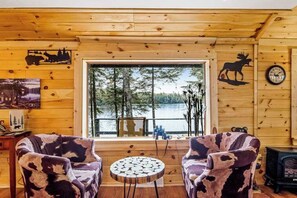 A cabin interior with wooden walls, two chairs with a cowhide pattern, a round wooden table, a large window with a view of trees and water, and a clock on the wall.