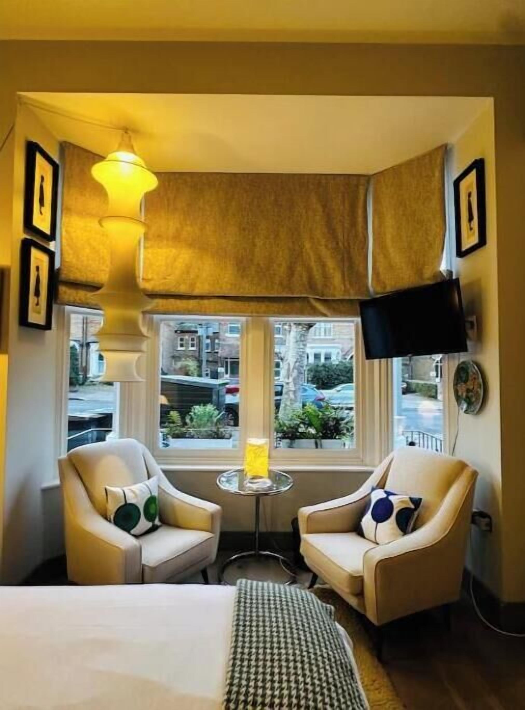 Habitación de hotel con cama, dos sillones, una mesita, una lámpara, televisor y ventana con vistas a edificios.