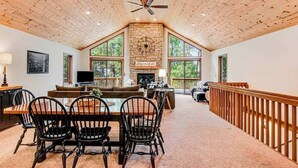 Un vaste espace de vie doté d’un haut plafond, d’une cheminée, d’une table à manger avec des chaises et de grandes fenêtres offrant une vue sur l’extérieur.