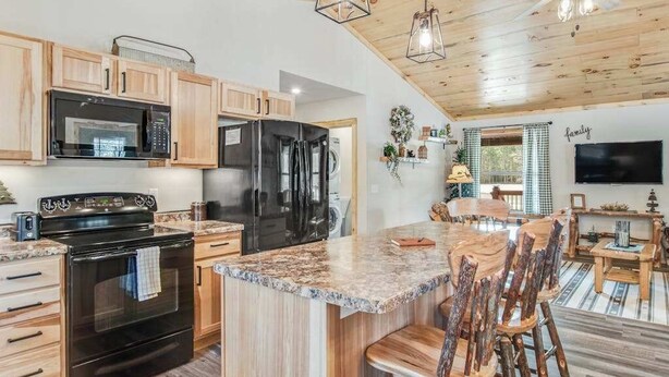 Cocina con armarios de madera, un refrigerador negro, un microondas y una estufa. Hay una zona de comedor con sillas y una mesa de madera.