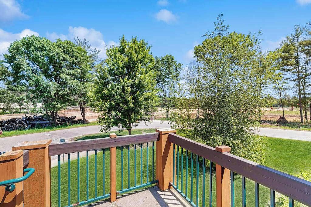 A wooden deck with a metal railing overlooking a grassy area with trees and a road.