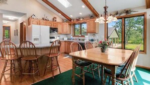 A modern kitchen with wooden cabinets, a dining area with a large table and chairs, and a view of the outdoors through large windows. - Golf course-front home with 2 pools, private beach, boat launches, tennis, grill (Nekoosa)