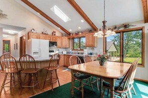 A modern kitchen with wooden cabinets, a dining area with a large table and chairs, and a view of the outdoors through large windows.