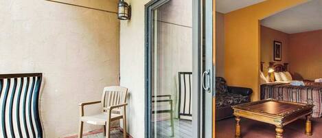Uma varanda com cadeira, mesa e porta de vidro deslizante que dá para um quarto com cama e sofá.