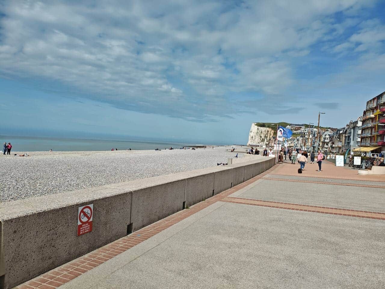 A coastal promenade with a pebble beach, a concrete barrier with a no swimming sign, and a row of colorful buildings in the background.