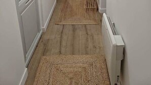 Een smalle gang met witte wanden, een houten vloer en een radiator. Links een deur en rechts een klein tafeltje met een mandje.