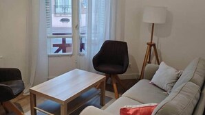 Een woonkamer met een bank, een houten salontafel, een zwarte stoel, een vloerlamp en uitzicht door een raam met gordijnen.