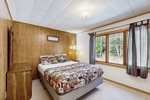 Une chambre avec un mur en bois, un lit avec une literie à motifs, une table de chevet et une fenêtre avec des rideaux.