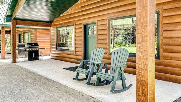 Une cabane en rondins dotée d’un patio couvert, de deux fauteuils Adirondack et d’un barbecue.