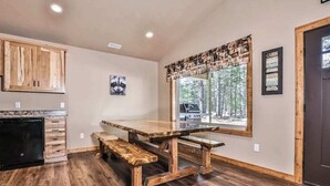 Une cuisine avec des meubles en bois, une table à manger avec des bancs, une fenêtre donnant sur la forêt et une porte portant l’inscription « CABANE ».