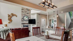 A modern living room with a fireplace, a dining area with a table set for four, and a kitchen in the background. - Newly-built condo on Duck Lake with decks, private dock, & firepit (Eagle River)