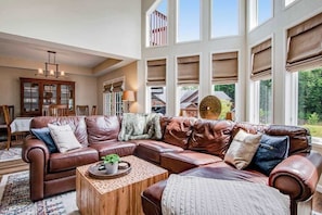 A spacious living room with a leather sectional sofa, a wooden coffee table, and large windows with blinds.