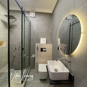 A modern bathroom with a glass shower enclosure, a marble sink, and a large mirror with a circular light. - Three Bedroom Luxury Apartment in Wuse 2 (Abuja)