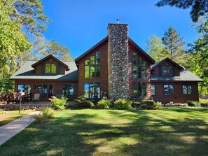 A two-story log cabin with a stone chimney, large windows, and a well-maintained lawn. - Lakefront home with game rooms, dock, kayaks, private beach, fireplace, firepit (Conover)